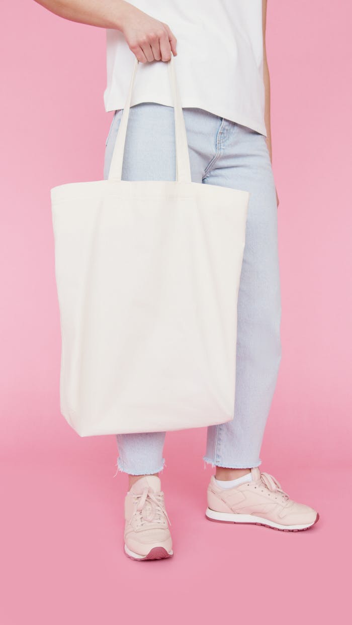Fashionable young woman holding a white tote bag against pastel pink background.